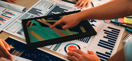 A diverse group of individuals engages in collaborative data analysis using a digital tablet, surrounded by printed charts and graphs in a modern workspace environment. SACTR