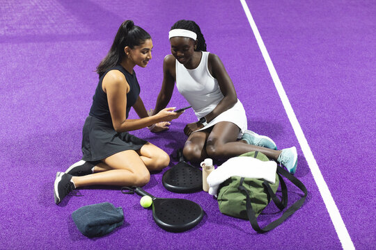 Diverse female friends sitting on purple court viewing smartphone with padel rackets, ball, bag