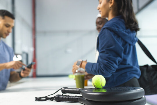 Diverse friends using smartphone and card and waiting at reception desk with padel tennis rackets