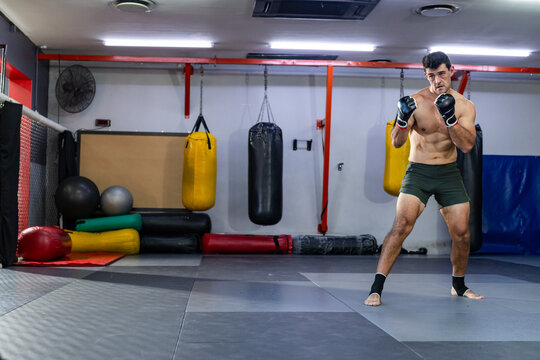 Shirtless male boxer punching bags on floor mats in boxing studio, copy space