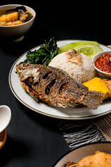 Whole fried fish with white rice, sambal, tempeh, tofu and vegetables on plate; Indonesian cuisine shot on dark background—perfect for menu, promo or recipe blog.