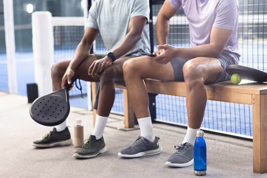 Diverse male friends sitting on wooden bench at padel court holding racket, checking smartphone