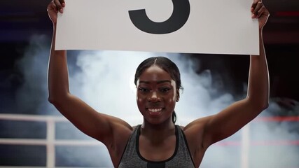young african american woman in crop top holds up sign with round number three in brightly lit stadium and smiles. athletic female at sports event with smoke in background. sportive concept, promotion - Powered by Adobe