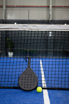 Padel racket leaning against net on blue court with white lines, yellow tennis ball resting