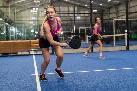 Female friends playing padel tennis on blue turf court under industrial lights with rackets