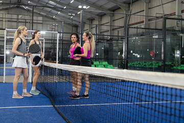 Diverse female padel players standing at net on blue-turf court in tank tops holding rackets
