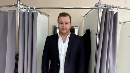 A Caucasian man in a tailored suit stands contemplatively in a fitting room, evoking the ritual of...