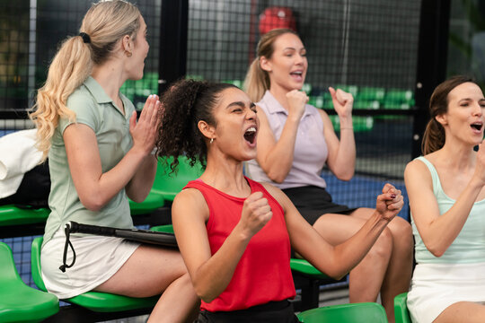 Female friends cheering on bleacher seats with bag strap at tennis court net behind fence