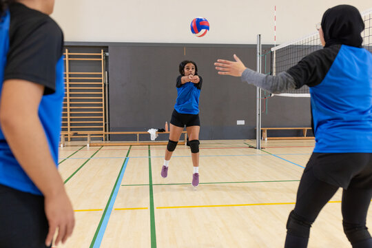 Diverse female team in blue jerseys bumping volleyball over net in gym with wall bars