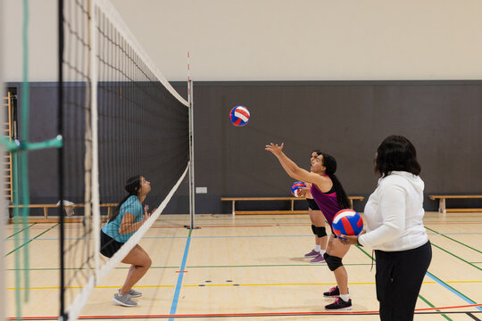 Diverse female teammates in sportswear practicing volleyball drills at gym with net, volleyballs