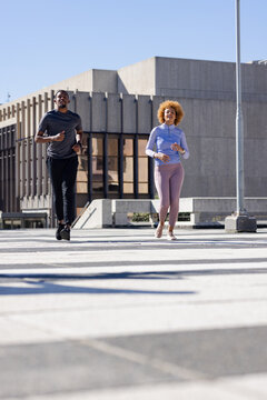 Diverse running partners jogging across crosswalk at plaza in sportswear near bench, lamp post