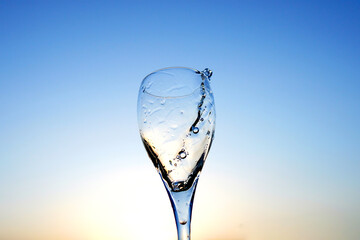 Champagne splashes from champagne glass on sunset blue sky. Romantic glass of white wine with bubbles, splashes. Champagne spilling from glass. Sparkling wine bubbles splash. Vibrant liquid splashing.