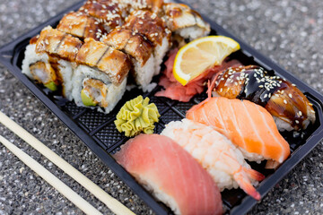 A diverse selection of sushi is arranged on a black tray, showcasing rolls topped with eel and lemon slices alongside vibrant nigiri and wasabi in daylight