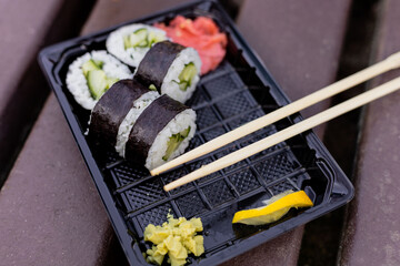 Delicious sushi rolls filled with cucumber are neatly arranged in a black tray alongside wasabi, pickled ginger, and a lemon slice on a rustic picnic table outdoors