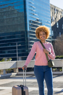 African american woman pulling suitcase in plaza wearing pink sweater, green crossbody bag