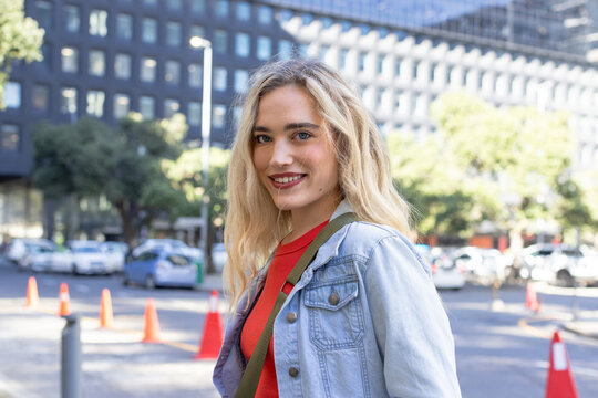 Woman standing on city street in denim jacket carrying green bag, traffic cones, copy space