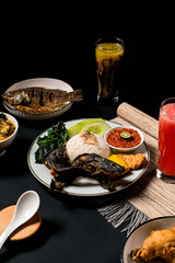Indonesian rice plate with fried catfish (nasi lele), sambal, tempeh, fried tofu, greens, cucumber, plus vegetable soup and juice on dark background—ideal for menu or promo.