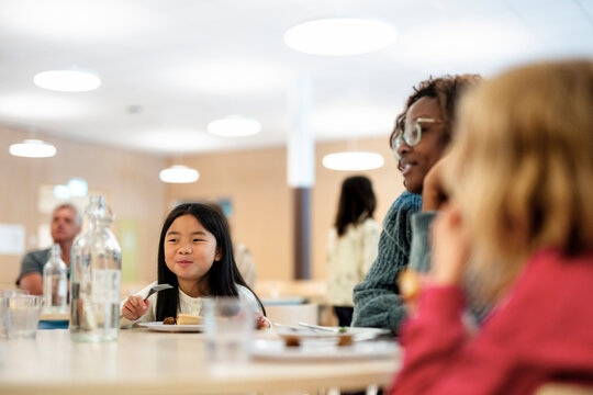 Smiling girl eating meal with teacher and friends while sitting in school cafeteria