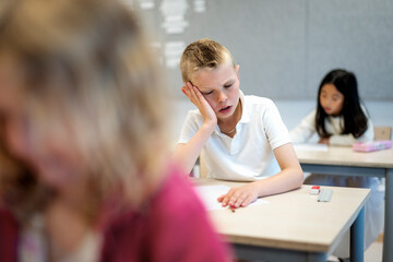 Tired boy leaning on elbow while sitting at desk in elementary classroom