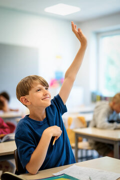 Smiling elementary boy raising hand while sitting at desk in classroom