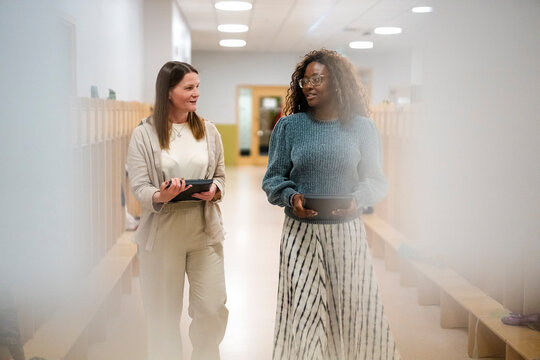 Female teachers communicating with each other holding laptops while walking in corridor