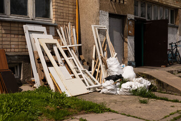 A collection of discarded doors, wooden frames, and garbage bags are stacked against a dilapidated building. Sunlight illuminates the scene, revealing overgrown grass and neglected surroundings