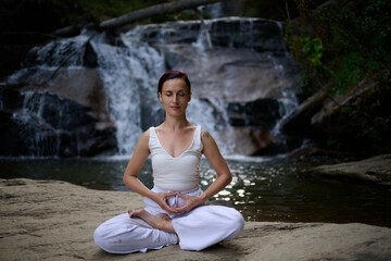Young woman sitting in lotus pose under tropical waterfall meditating with calm strength and mindfulness fully connected to nature energy tranquility and inner balance