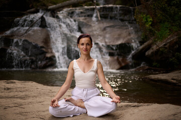 Young woman sitting in lotus pose under tropical waterfall meditating with calm strength and mindfulness fully connected to nature energy tranquility and inner balance