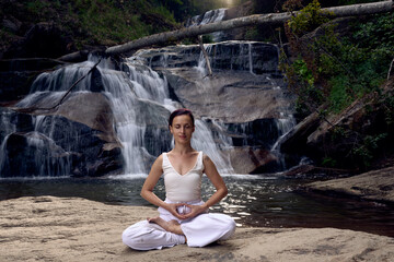 Young woman sitting in lotus pose under tropical waterfall meditating with calm strength and mindfulness fully connected to nature energy tranquility and inner balance