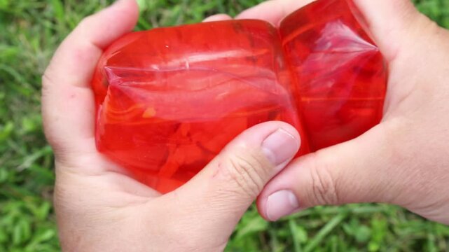 Hands hold and squeeze the translucent red anti -strats in the open air on green grass, elastic surfaces are dynamically deformed under pressure in daylight. ASMR
