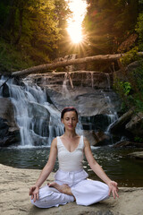 Young woman sitting in lotus pose under tropical waterfall meditating with calm strength and mindfulness fully connected to nature energy tranquility and inner balance