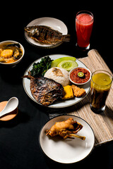 Indonesian rice plate with whole grilled fish, sambal, tempeh, fried tofu, greens, and cucumber, plus juice on the side. Vertical, moody food styling for restaurant promotion.
