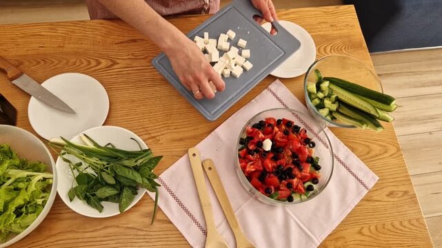 A girl puts sliced feta cheese into a salad bowl. A girl in an apron prepares a healthy vegetable salad. A bowl of vegetables stands on the table. Preparing a Greek salad. Top view