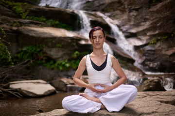 Young woman sitting in lotus pose under tropical waterfall meditating with calm strength and mindfulness fully connected to nature energy tranquility and inner balance