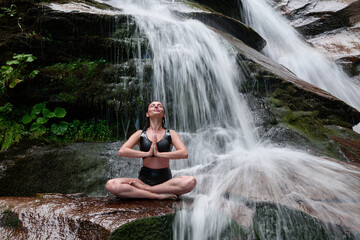 Young woman sitting in lotus pose under tropical waterfall meditating with calm strength and mindfulness fully connected to nature energy tranquility and inner balance