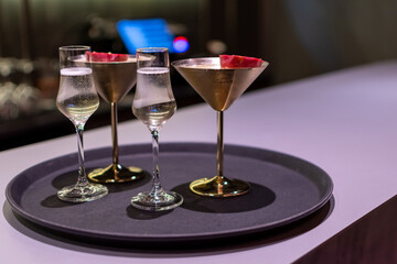Elegant cocktails, including two clear sparkling drinks and two gold-colored Porn star martinis with red garnishes on dark circular tray on background of blurred bar scene