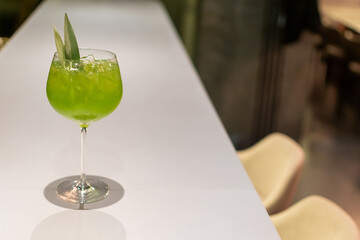 Tall glass with bright green cocktail Basil Smash and leaf garnish alone on clean, light-colored bar top on dark background