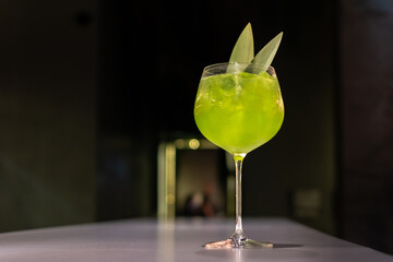 Vibrant green cocktail Basil smash in large glass with ice and garnish of two leaves on bar with dark blurred background