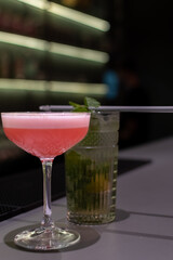 Vibrant pink cocktail with light foam top is served in coupe glass, placed on black bar mat next to mojito. The background is dark and out of focus
