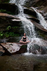 Young woman sitting in lotus pose under tropical waterfall meditating with calm strength and mindfulness fully connected to nature energy tranquility and inner balance