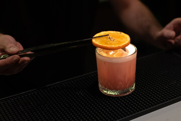 Bartender is using tongs to place a torched orange slice on top of a finished cocktail in a glass