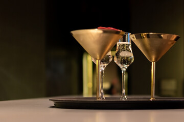 Porn star martini cocktail. Two unique gold glasses and clear, elegant glass with sparkling wine on black serving tray. The background is dark and blurred