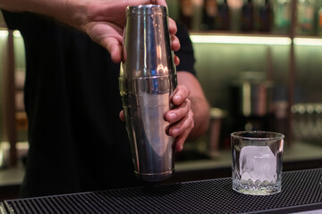 Bartender stirring and shaking the drink. A glass with a large ice cube is also visible