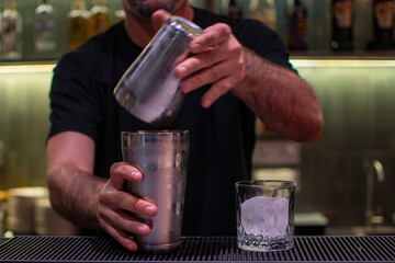 Bartender is expertly separating the two parts of a metal cocktail shaker, with a glass of ice ready on the bar