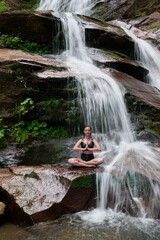 Young woman sitting in lotus pose under tropical waterfall meditating with calm strength and mindfulness fully connected to nature energy tranquility and inner balance