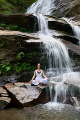 Young woman sitting in lotus pose under tropical waterfall meditating with calm strength and mindfulness fully connected to nature energy tranquility and inner balance