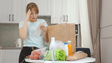 Young woman standing in kitchen with worried expression, checking grocery receipt and experiencing shock from high food prices, reflecting economic challenges of rising living costs