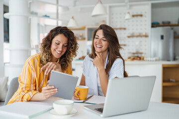 Two Female Friends Using Laptop And Tablet During Remote Work Meeting In Bright Contemporary Kitchen Space