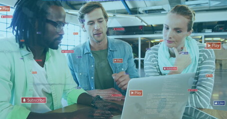 Collaborating team reviewing laptop screen around table in loft office, floating digital icons