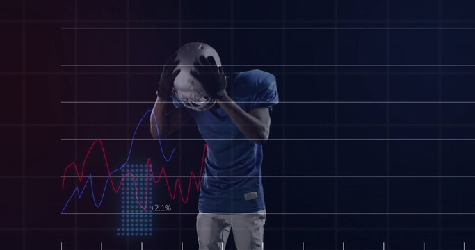 Clutching helmet to head, football player wearing blue jersey standing in studio, with data graphs